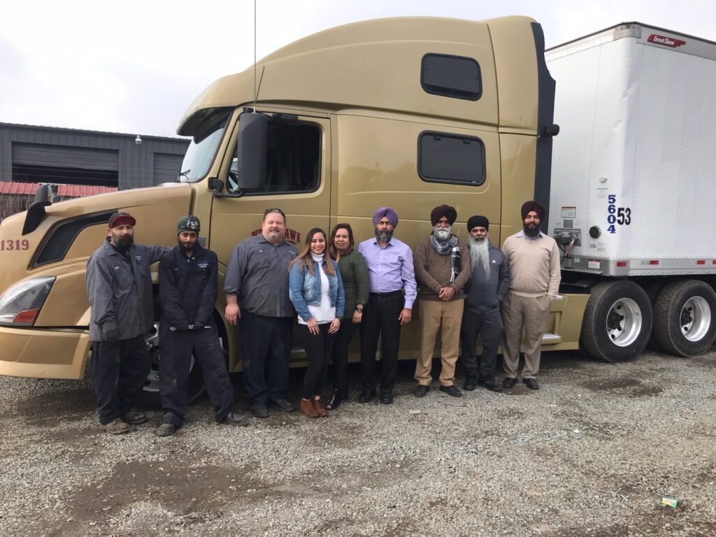 Eight people stand side by side in front of a beige semi-truck and trailer on a gravel lot, posing for a group photo—some in turbans and work uniforms, others in business casual attire, ready to tackle the busy Retail Calendar ahead.