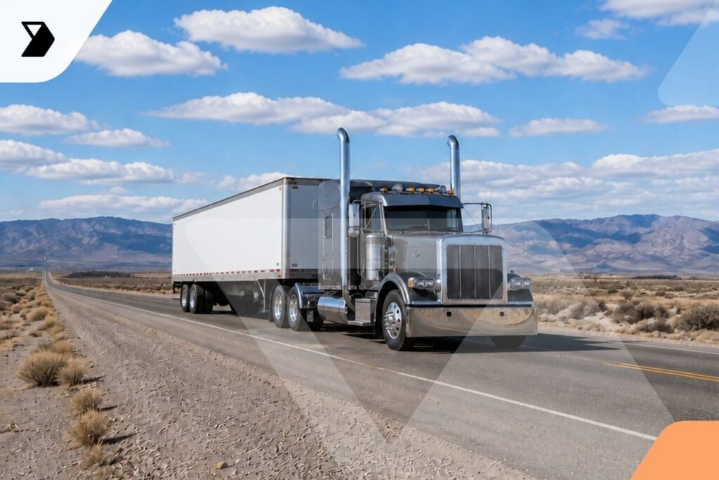 A large silver semi-truck with a white trailer drives down an empty desert highway under a partly cloudy sky, with mountains visible in the background.