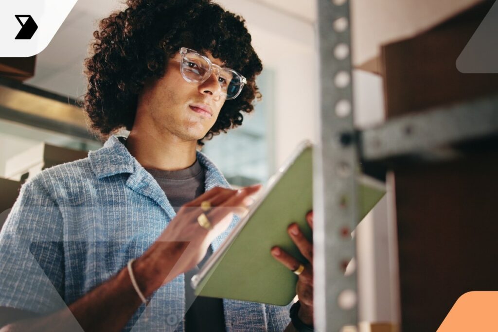 A person with curly hair and glasses uses a tablet in a workspace, possibly taking inventory or notes.