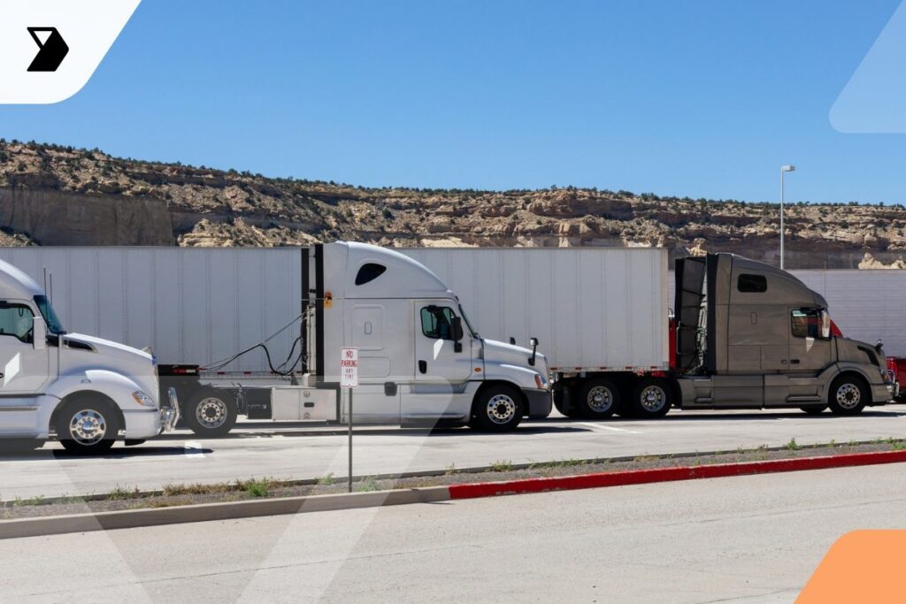 Several semi-trucks are parked side by side in a rest area under a clear blue sky, with rocky hills in the background and red curbing along the pavement.