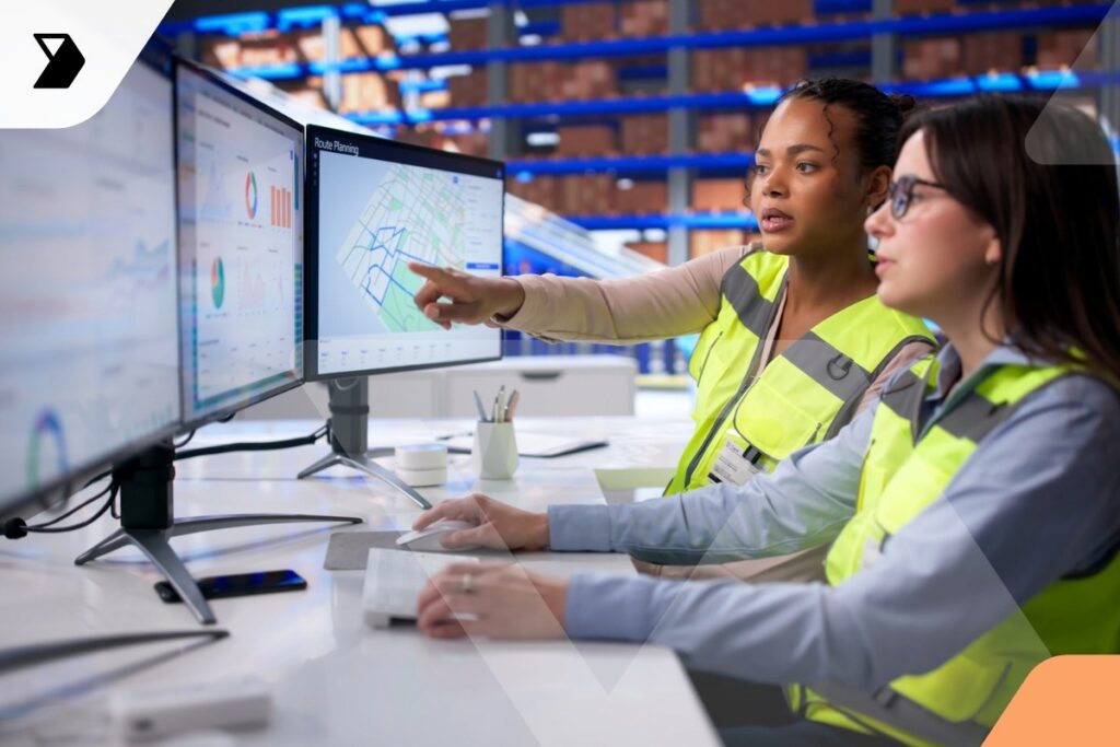 Two women wearing safety vests sit at a desk with multiple monitors displaying charts, maps, and Shipping KPIs.
