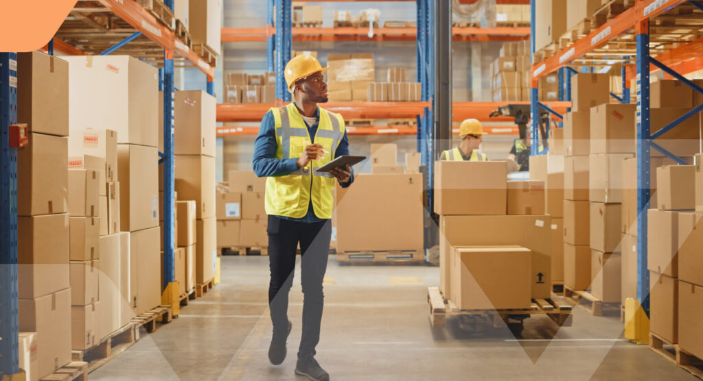 Warehouse manager in a hard hat and safety vest uses a tablet while walking through an aisle of stacked boxes