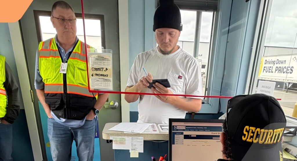A man in a safety vest and another man in a beanie stand at a counter; one uses a phone. A person wearing a SECURITY hat sits at a computer in the foreground. Signs about fuel prices and paperwork are visible.