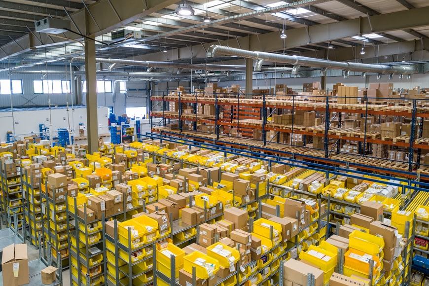 A large warehouse filled with shelves stacked with numerous yellow bins and cardboard boxes, organized and well-lit with industrial lighting and metal beams visible above.