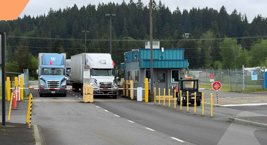 Two semi-trucks and a small utility vehicle wait at a security checkpoint booth surrounded by yellow poles, with a forested area in the background—an efficient scene where Bill of Lading Management cuts losses and streamlines cargo processing.