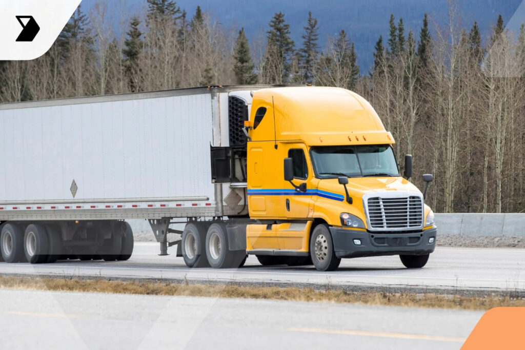 A bright yellow semi-truck with a white trailer drives on a highway, with trees and mountains in the background