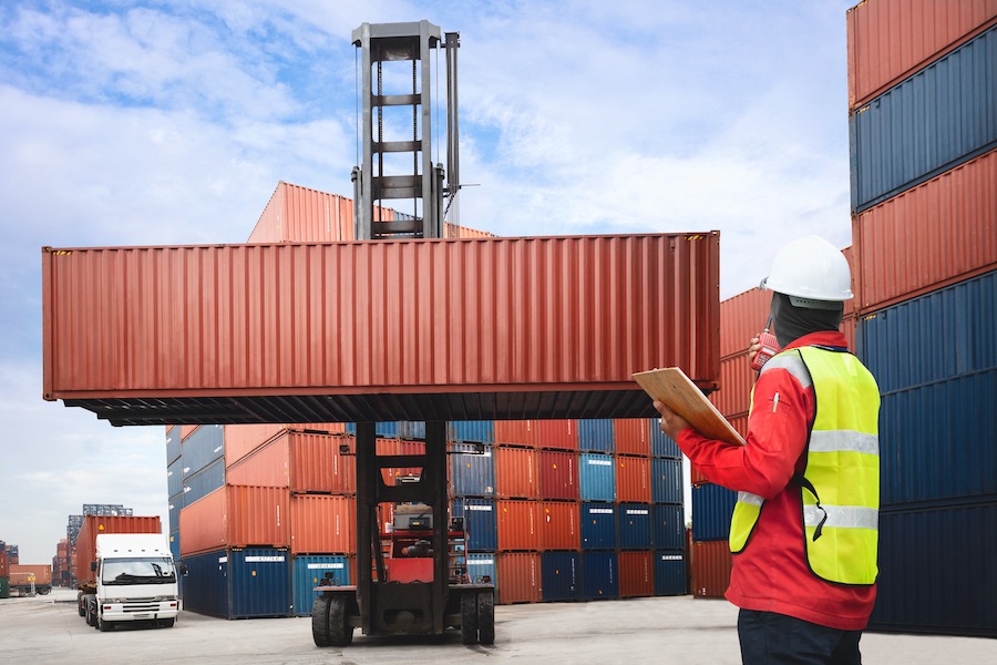 A worker in a safety vest and helmet holds a clipboard and observes as a forklift lifts a large shipping container in a busy shipping yard filled with stacked containers.