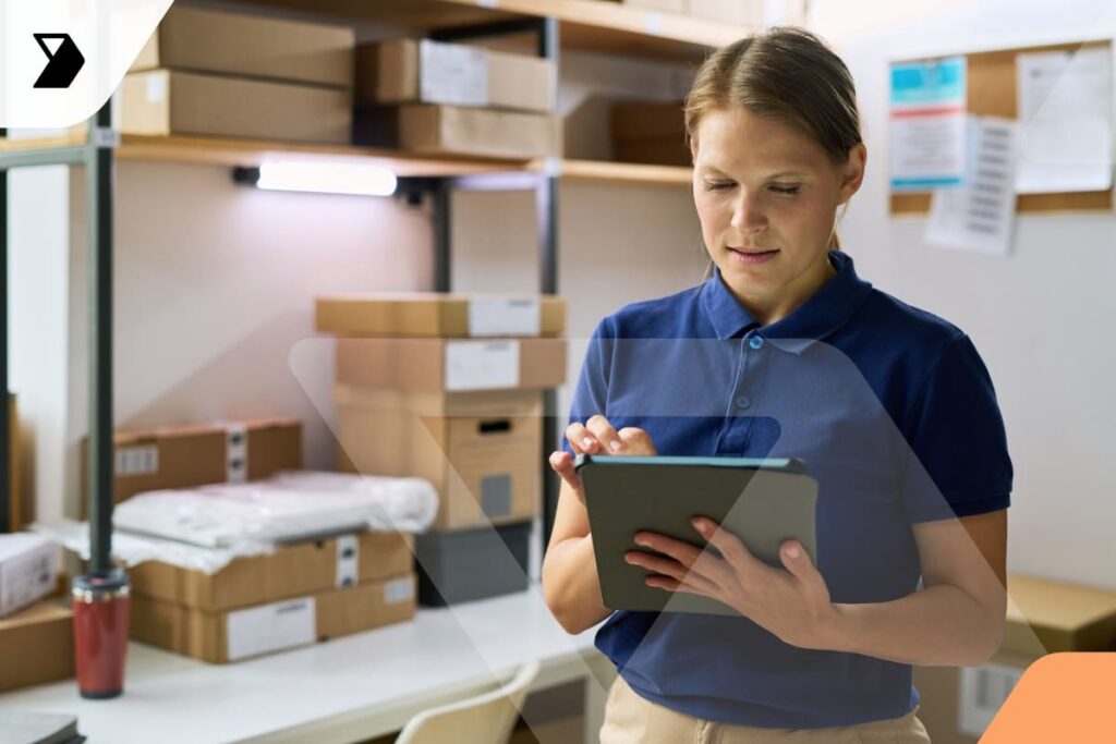 A woman in a blue polo shirt uses a tablet in a room with shelves filled with cardboard boxes, likely managing inventory or handling proof of delivery in logistics.