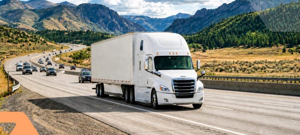 A white semi-truck drives on a multi-lane highway surrounded by mountains and green hills under a partly cloudy sky, with several cars visible in the opposite lane.