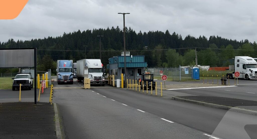 Two semi-trucks stop at a small blue and white inspection booth with yellow poles and stop signs, surrounded by forest and cloudy sky. Several other vehicles and a portable toilet are nearby.