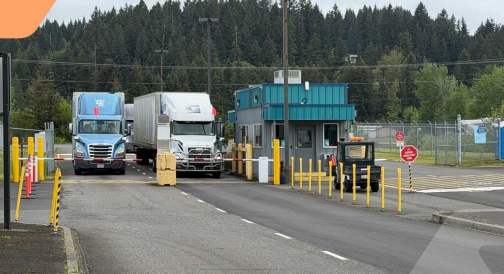 Two semi-trucks stop at a security checkpoint with a small teal booth. Yellow barriers and bollards line the road. Trees and a fence are visible in the background under an overcast sky.