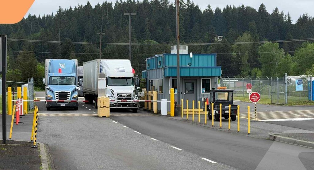 Two semi-trucks are stopped at a security checkpoint booth on a paved road surrounded by trees and fencing, with yellow poles and stop signs visible.