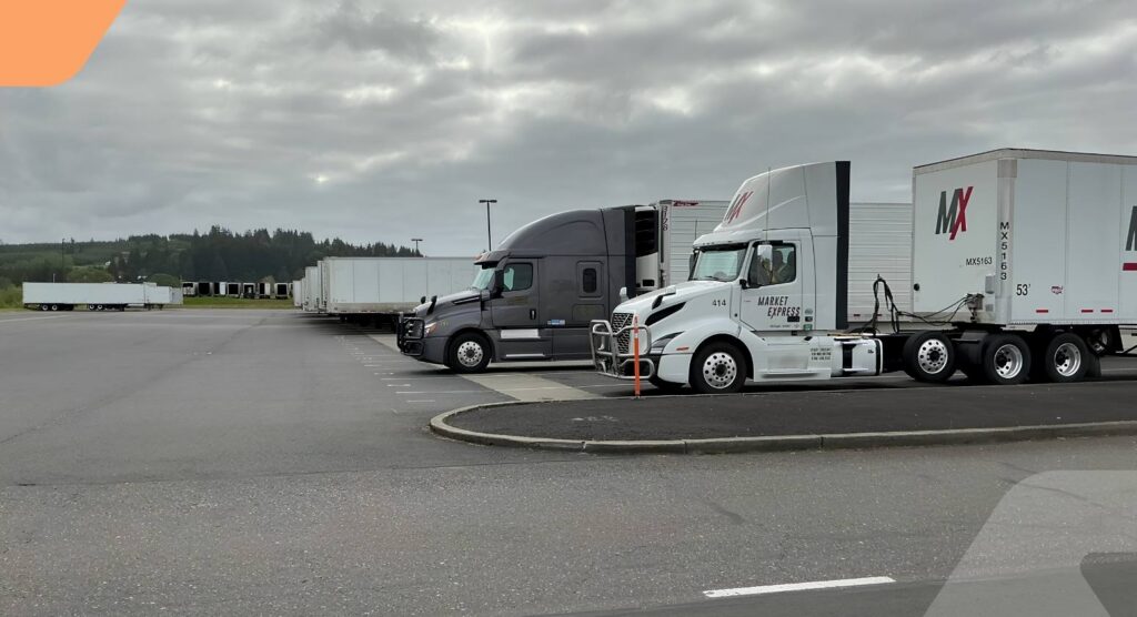 Several semi-trucks are parked in a mostly empty, paved lot under a cloudy sky, with trees and grassy areas visible in the background.